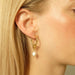 Close-up of a woman adorned with the Catherine Earrings by Noémiah, showcasing elegant gold-plated hoops with freshwater pearls. Her blonde hair is tucked behind her ear, enhancing her light, translucent green top against a neutral background.