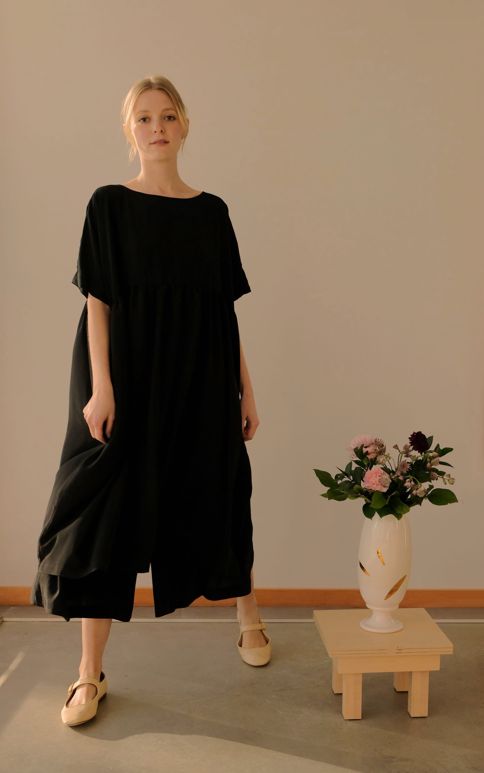 A person in the Cécilia Classic Dress by Noémiah, made from elegant black silk and featuring 3/4 dolman sleeves, stands indoors beside a small pedestal holding a vase with assorted flowers. The room has a minimalist aesthetic with neutral tones.