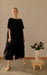 A person in the Cécilia Classic Dress by Noémiah, made from elegant black silk and featuring 3/4 dolman sleeves, stands indoors beside a small pedestal holding a vase with assorted flowers. The room has a minimalist aesthetic with neutral tones.