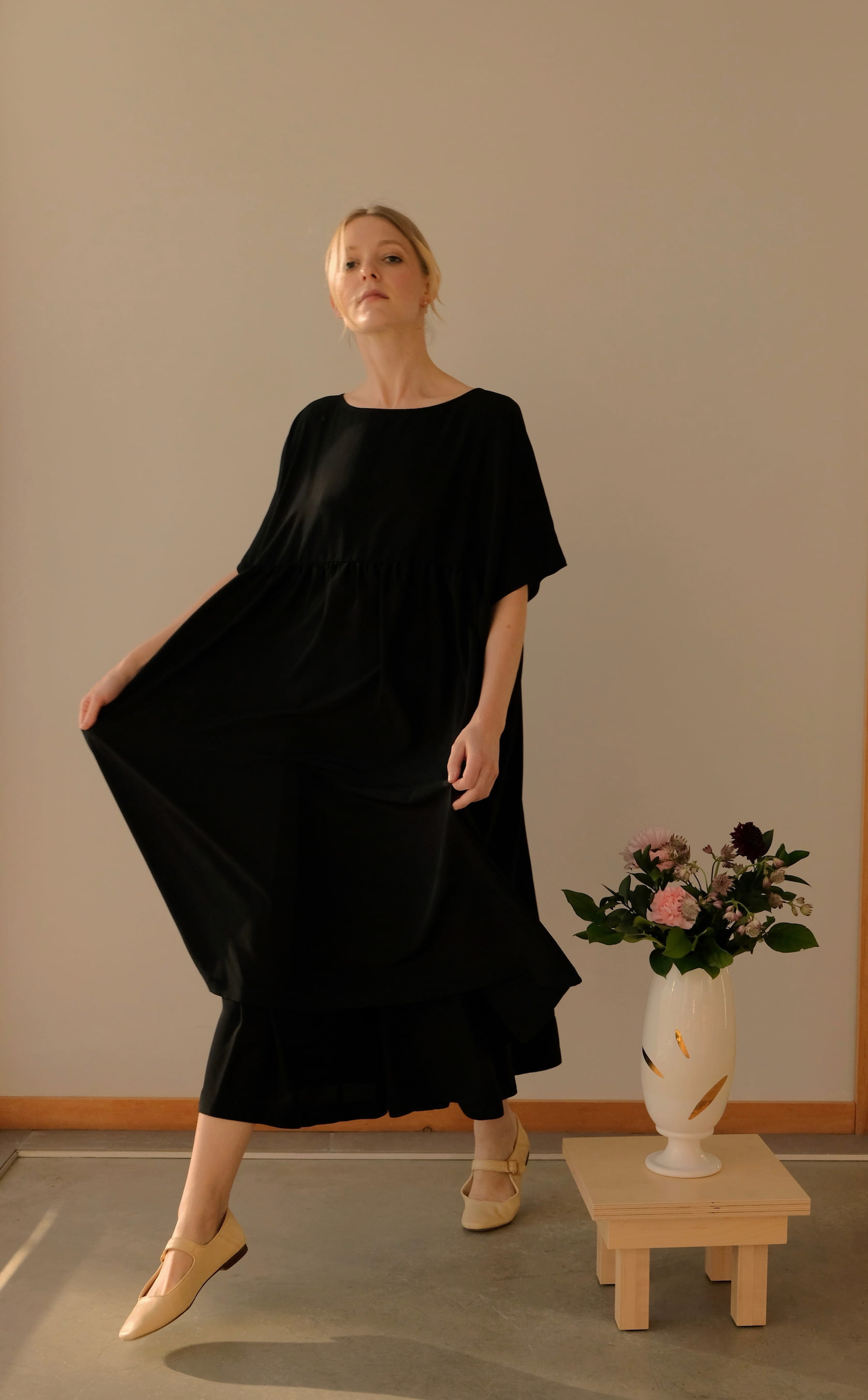 A person stands holding the hem of the Cécilia Classic Dress in black silk, featuring 3/4 dolman sleeves. They are in a room with a light gray wall and wooden flooring. Next to them is a white vase with pink and dark purple flowers placed on a wooden stool. The dress is from the brand Noémiah.