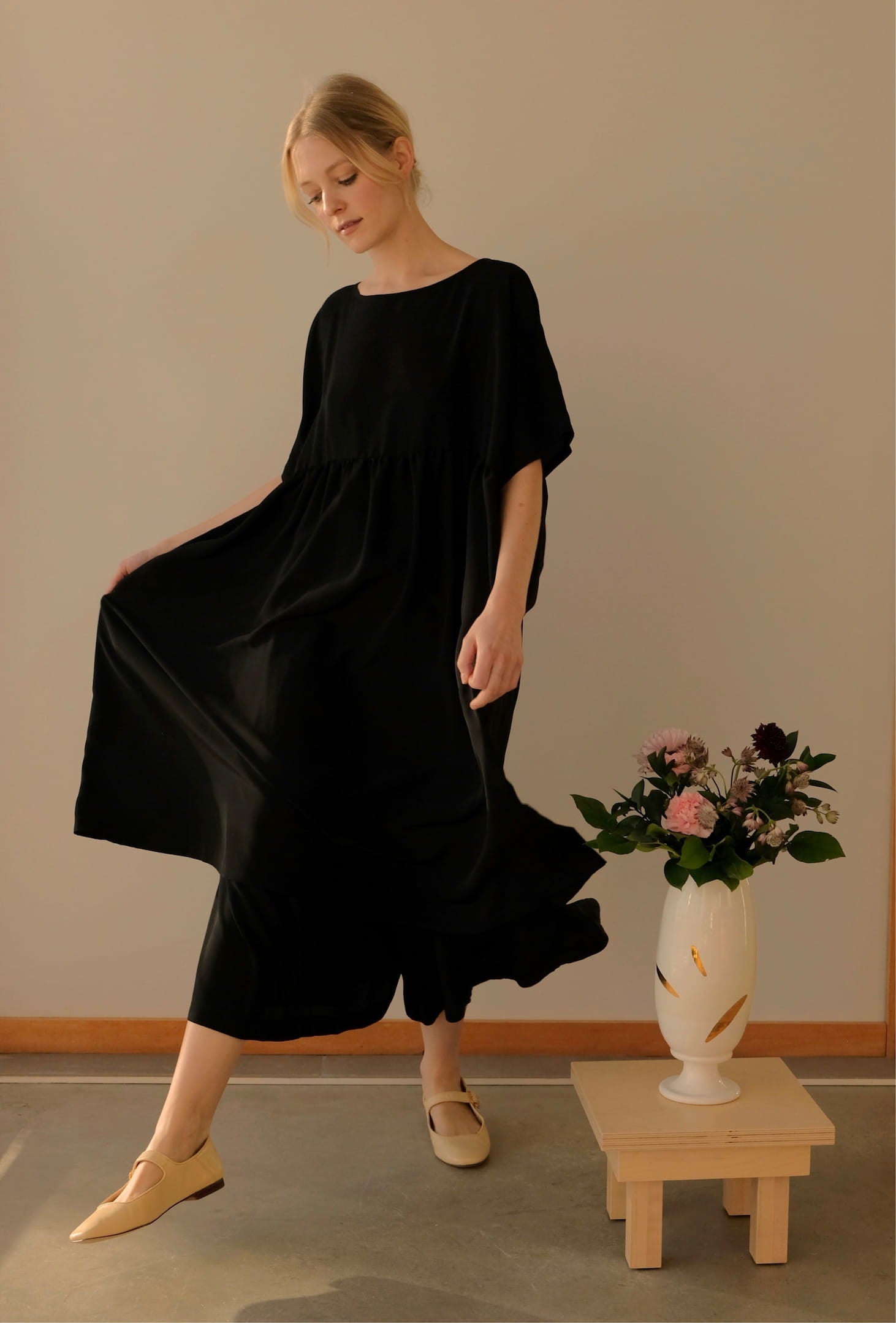 A person wearing the Cécilia Classic Dress in black silk by Noémiah, featuring 3/4 dolman sleeves, poses gracefully indoors, accompanied by beige shoes and standing beside a wooden stand with a white vase filled with assorted flowers. The background is a simple, neutral-colored wall.