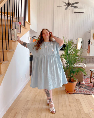 A person wearing a blue Cécilia Classic Dress by Noémiah and white sandals stands in a stylish living room next to wooden stairs. The room, adorned with a plant, sofa, and wall art, exudes a cozy atmosphere. They have tattoos and are smiling, with one hand resting on the staircase.