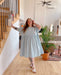 A person wearing a blue Cécilia Classic Dress by Noémiah and white sandals stands in a stylish living room next to wooden stairs. The room, adorned with a plant, sofa, and wall art, exudes a cozy atmosphere. They have tattoos and are smiling, with one hand resting on the staircase.