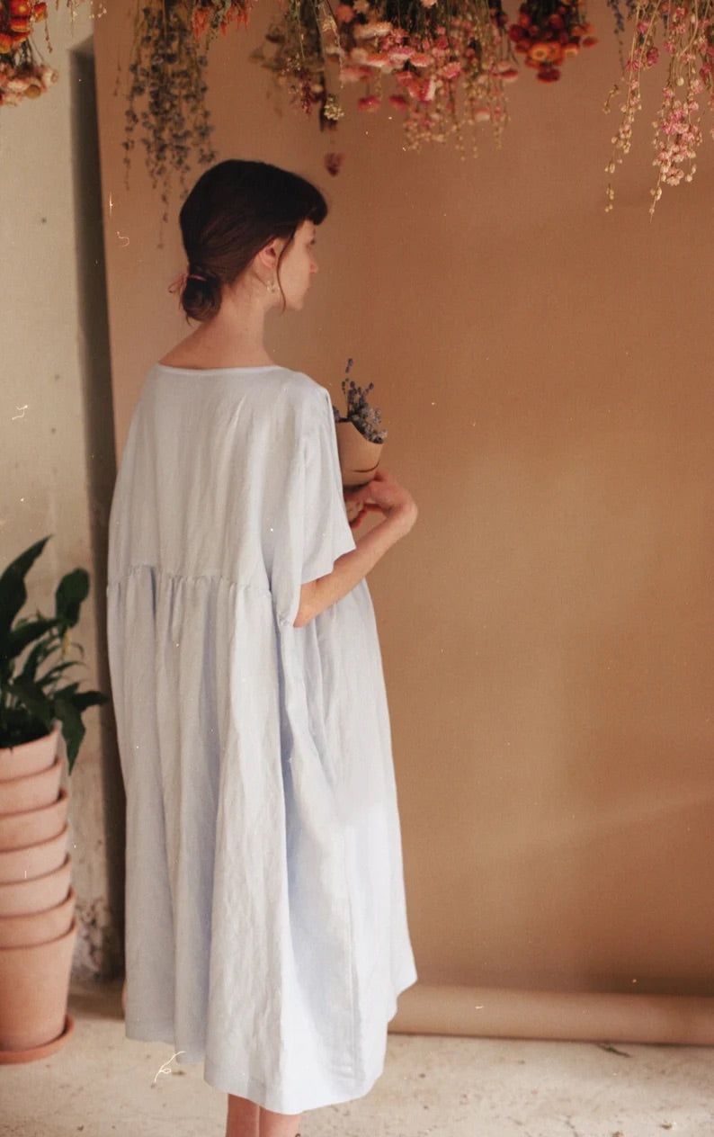 A woman wearing the Cécilia Classic Dress in blue by Noémiah stands holding dried lavender in a cozy room. The background features hanging flowers and several potted plants, with embroidered daisies enhancing the warm, natural ambiance.