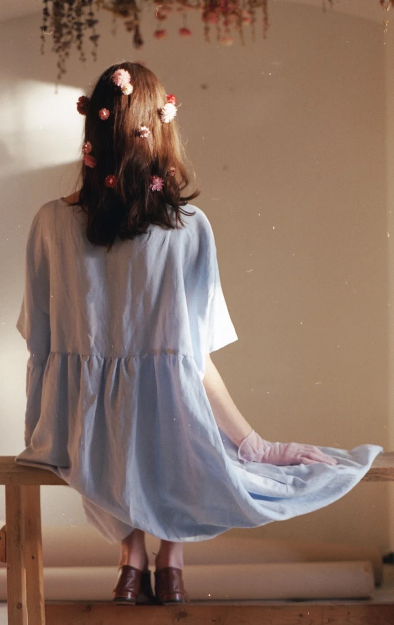 A person with long hair sits on a wooden bench, facing away, wearing a light blue Cécilia Classic Dress from Noémiah, adorned with pink flowers in their hair. Beige walls and hanging dried flowers create a warm, serene atmosphere filled with delicate embroidered daisies.