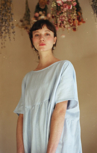 A person with short dark hair wearing a light blue Noémiah Cécilia Classic Dress stands in a softly lit room. Hanging flowers adorn the background, and embroidered daisies on the linen blend fabric enhance the warm, natural ambiance.