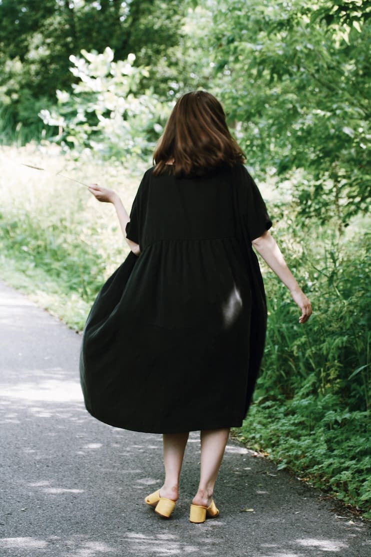 A woman in a Cécilia Classic Dress by Noémiah, crafted from breathable lyocell, and wearing yellow shoes strolls along a shaded path enveloped by lush green foliage, with her back turned towards the camera. In one hand, she holds a slender stick.