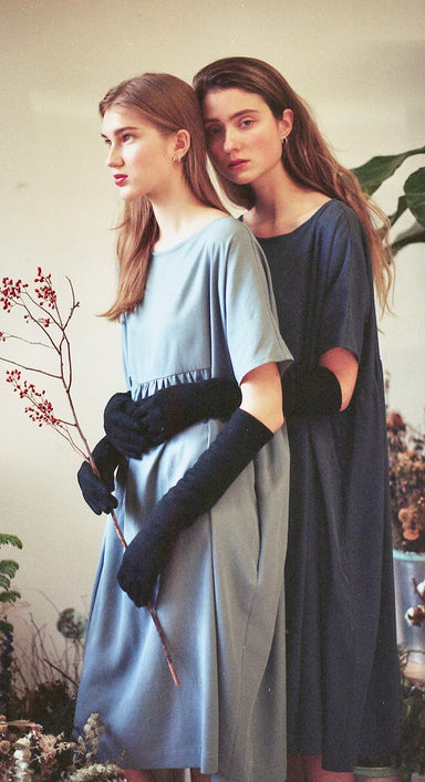 Two women stand closely, donned in Cécilia Classic Dresses made from Lyocell by Noémiah, paired with long black gloves. One gently holds a twig adorned with small red berries, capturing the spirit of sustainable fashion. Their introspective expressions are set against a neutral background accented with foliage.