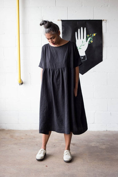 A person with grey hair stands in a Noémiah Cécilia Classic Dress made of lyocell fabric, paired with white shoes. They are looking down in front of a white brick wall that features a black banner adorned with a white hand and small yellow flowers.