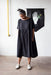 A person with grey hair stands in a Noémiah Cécilia Classic Dress made of lyocell fabric, paired with white shoes. They are looking down in front of a white brick wall that features a black banner adorned with a white hand and small yellow flowers.