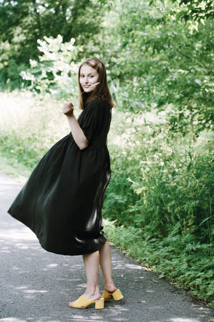 A woman in the Cécilia Classic Dress by Noémiah, crafted from breathable Lyocell fabric, and wearing yellow sandals twirls along a path enveloped by lush greenery. The sun filters through the leaves, creating a vibrant, summery scene that celebrates sustainable beauty.