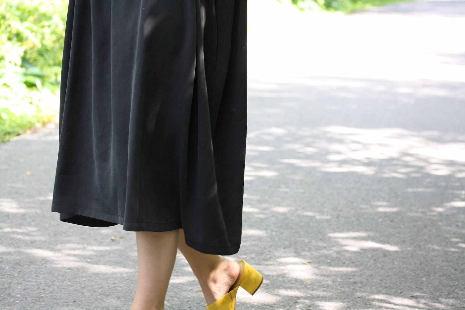 A person, clad in the Cécilia Classic Dress by Noémiah and yellow shoes, walks along a sun-dappled path flanked by vibrant greenery. The dress, made from breathable, sustainable lyocell, harmonizes with the shaded ground. The photograph captures only their lower half as they embrace an eco-conscious journey.