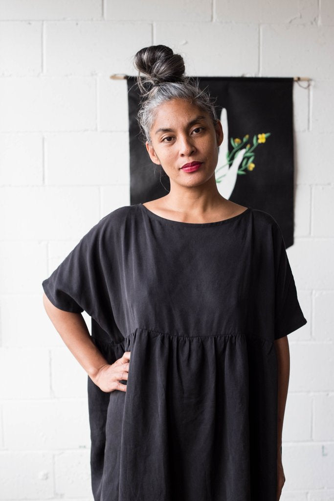 A woman with grey hair in a bun stands confidently in front of a white brick wall, embellished with a black banner featuring a simple design. She is dressed in the Cécilia Classic Dress from Noémiah, made of breathable lyocell fabric, and has one hand on her hip, exuding a relaxed yet poised demeanor.