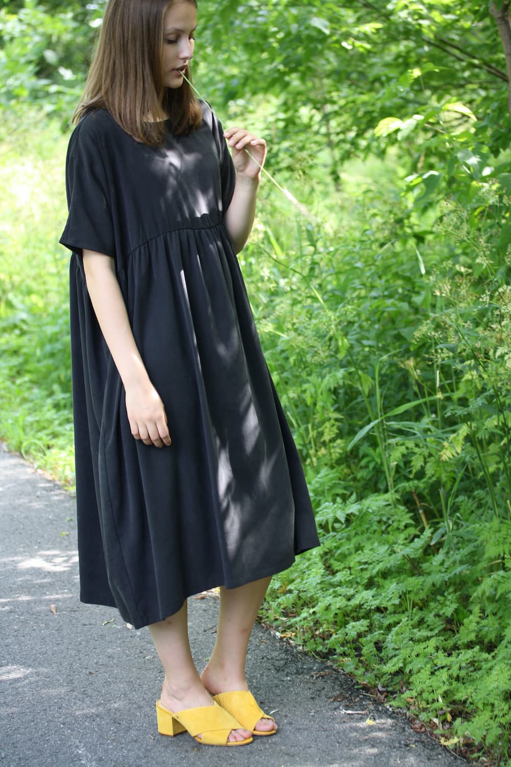 A person clad in the Cécilia Classic Dress by Noémiah, crafted from dark lyocell, pairs it with yellow shoes and stands thoughtfully on a path encircled by lush green foliage.