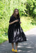 A woman stands on a sunlit path surrounded by lush greenery, wearing the loose, breathable Cécilia Classic Dress in black lyocell by Noémiah and a pair of yellow shoes. She holds a plant stem in one hand as shadows from nearby trees create patterns on the ground and her dress.