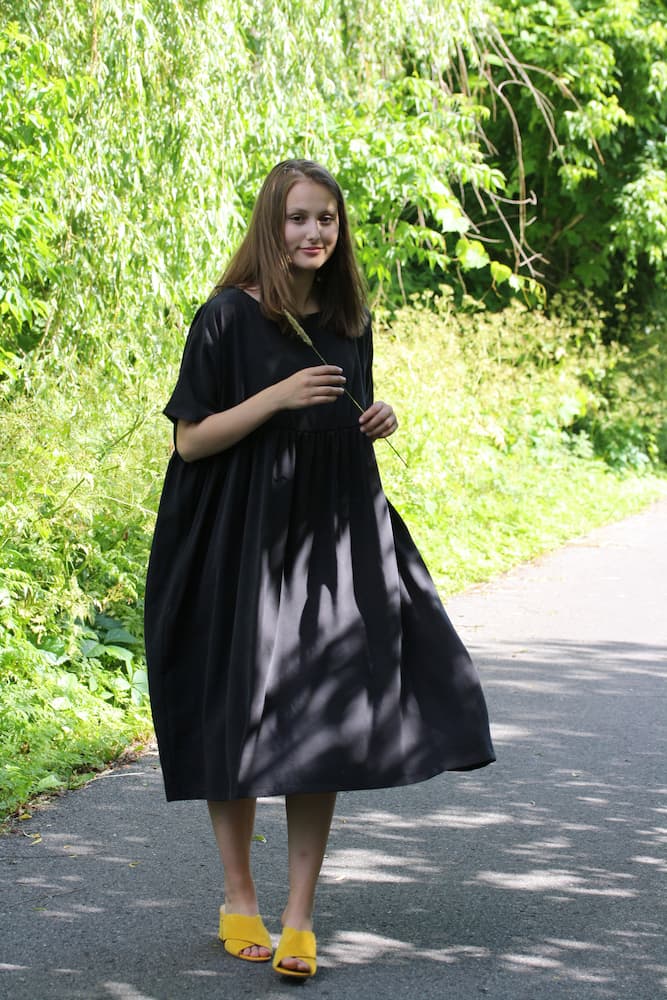 A woman stands on a sunlit path surrounded by lush greenery, wearing the loose, breathable Cécilia Classic Dress in black lyocell by Noémiah and a pair of yellow shoes. She holds a plant stem in one hand as shadows from nearby trees create patterns on the ground and her dress.