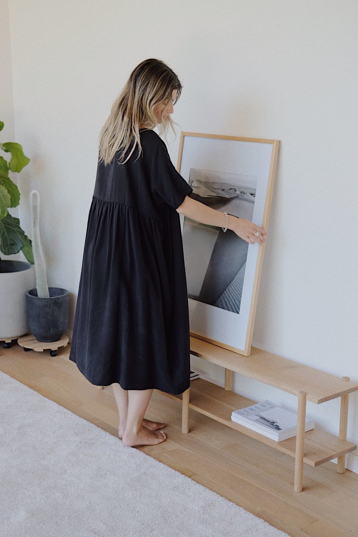 Wearing the Noémiah Cécilia Classic Dress in black lyocell, a woman places a framed artwork on a wooden bench in a minimalist room. To her left stands a large potted plant, and there is also a small stack of books on the bench. The room's sustainable design includes light-colored walls and a wooden floor.