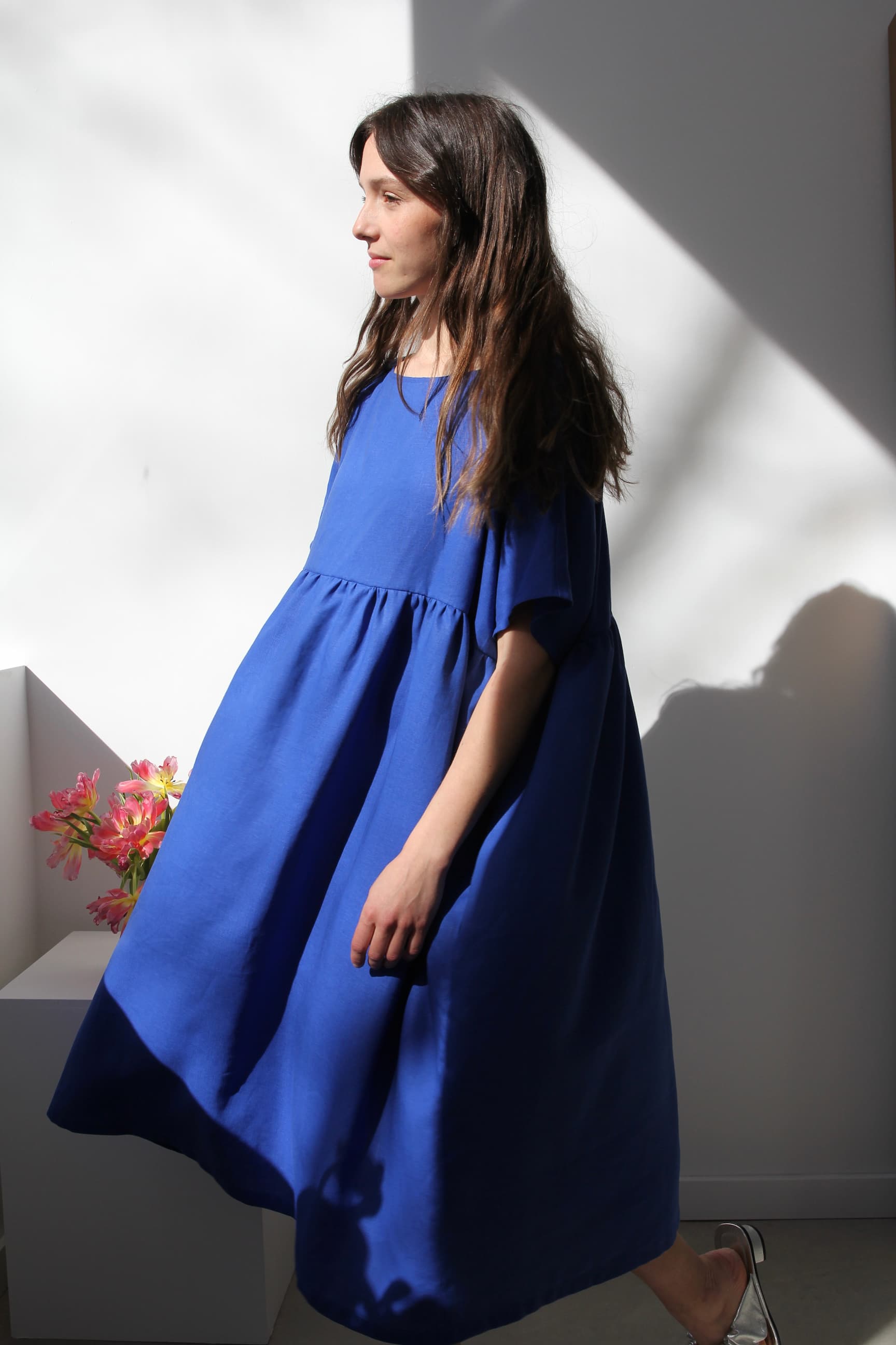 A woman wearing a relaxed, Matisse Blue Cécilia Classic Dress by Noémiah stands in a sunlit room. Pink flowers are displayed in a vase on a white shelf, embodying relaxed femininity as light and shadows dance around the scene.