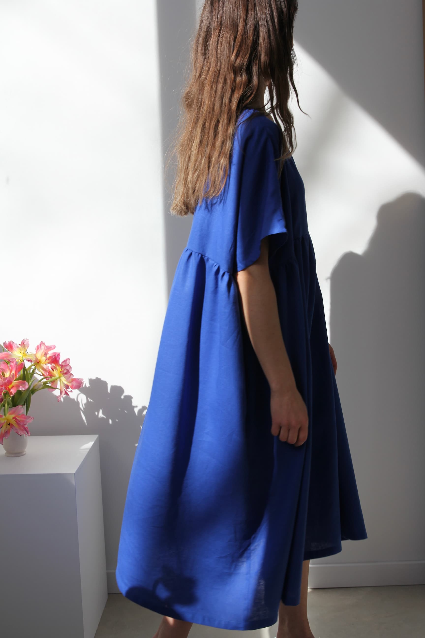 A woman with long hair stands in a room wearing the relaxed femininity of the Cécilia Classic Dress in Matisse Blue by Noémiah. She faces a white wall, where sunlight casts gentle shadows. On the left, a white table holds a vase of pink flowers, adding charm to the serene setting.