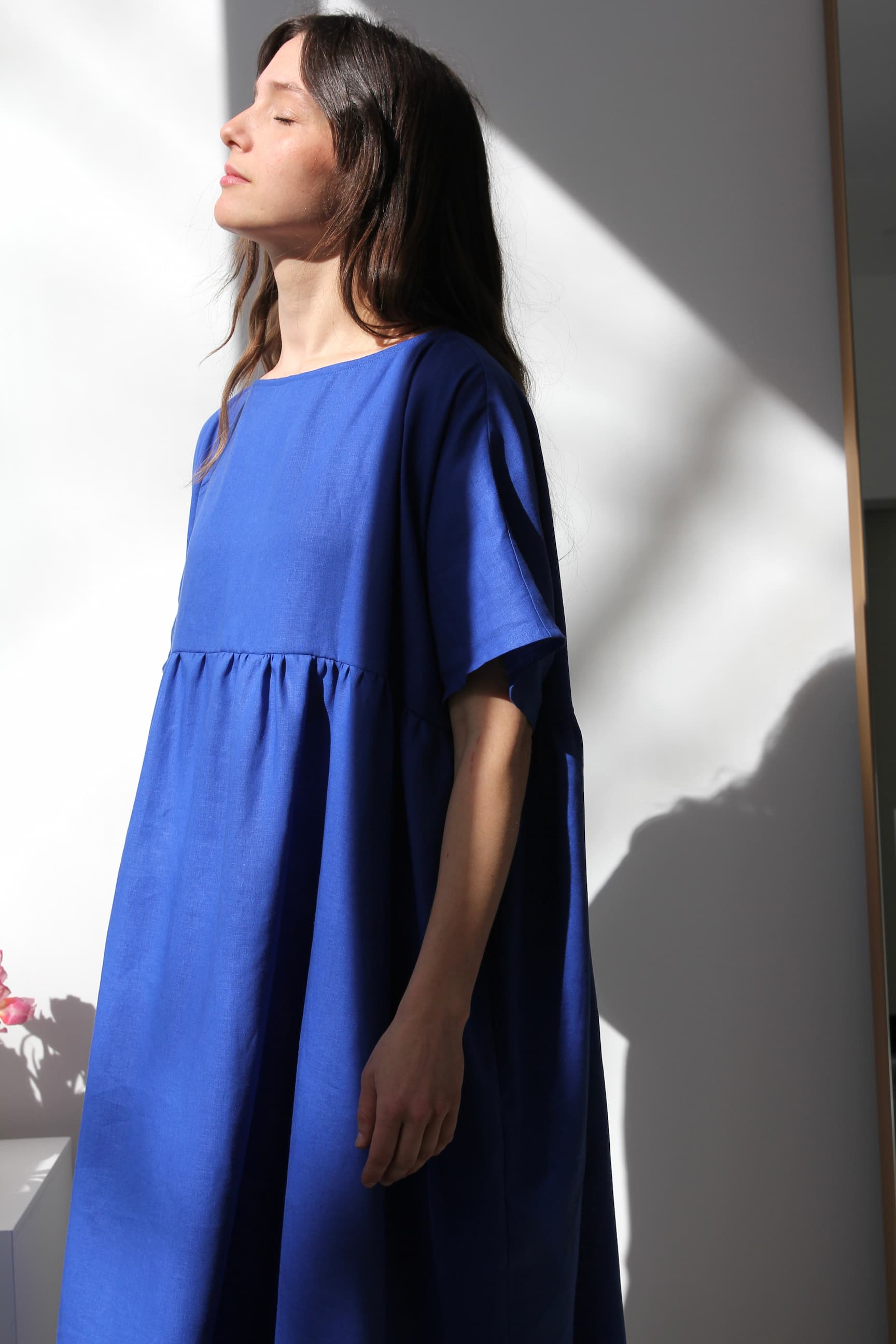 A woman stands in a flowing Cécilia Classic Dress — Matisse Blue by Noémiah, embodying relaxed femininity with her eyes closed as sunlight casts shadows on the wall, creating a calm and serene setting.