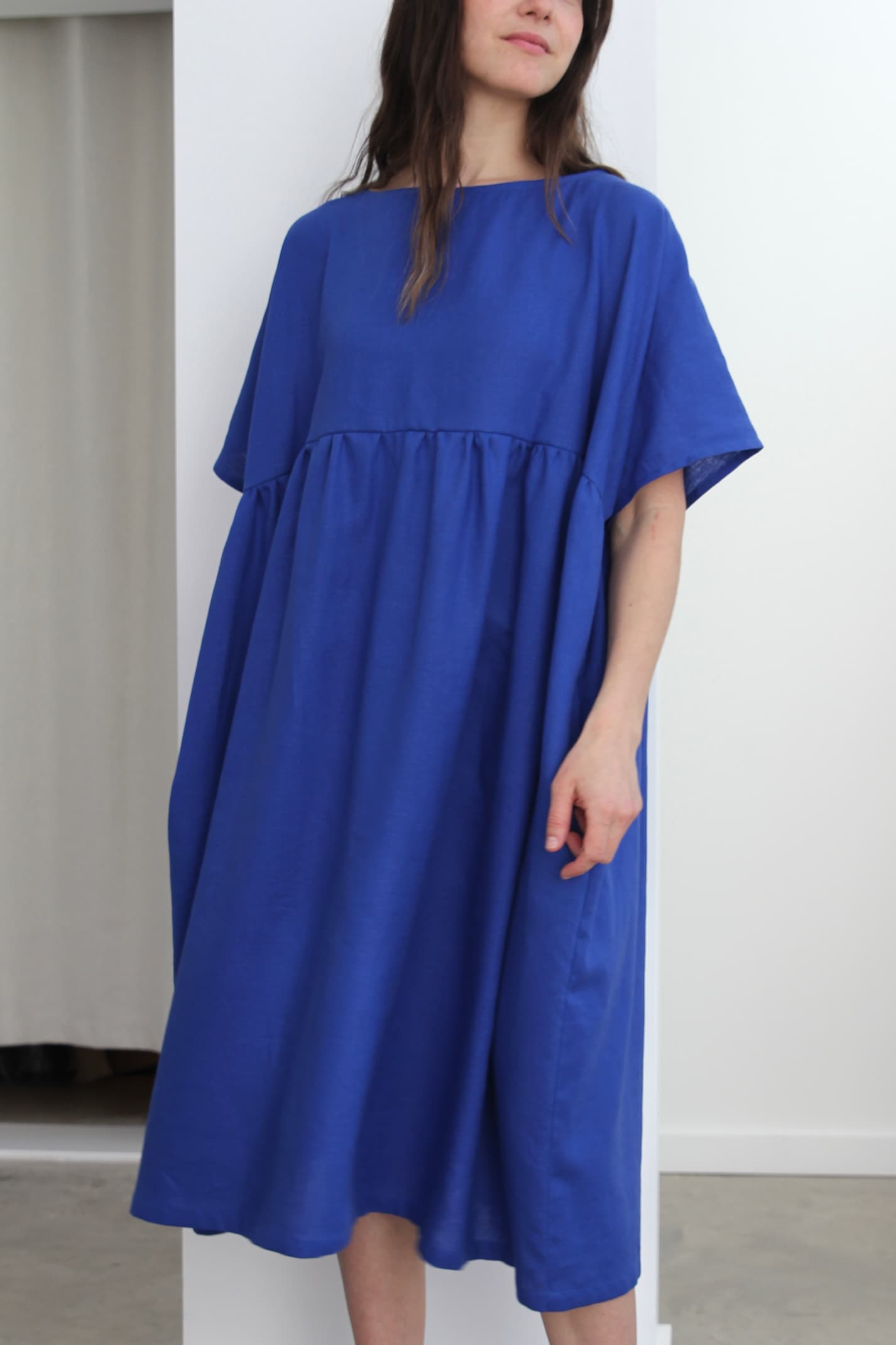 A person wearing a relaxed-fit Cécilia Classic Dress in Matisse Blue by Noémiah stands indoors against a white wall. With its short sleeves and gathered waist, this hand-wrapped piece exudes relaxed femininity and beautifully complements the minimalistic and modern setting.