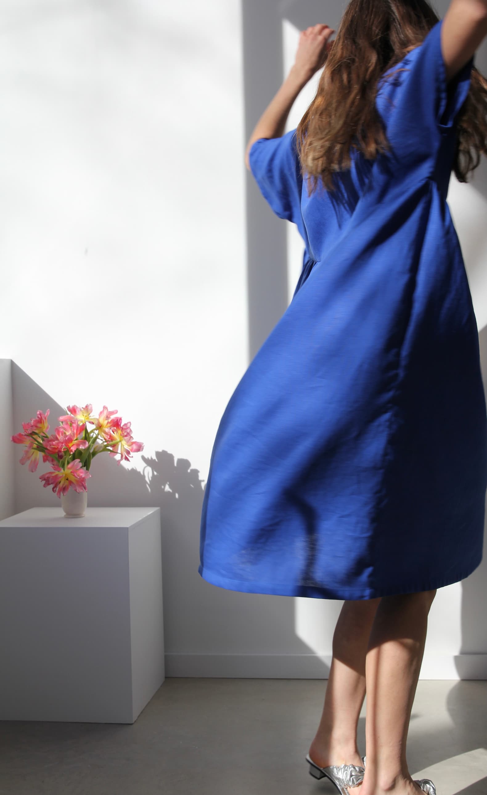 Dressed in the flowing Noémiah Cécilia Classic Dress in Matisse Blue, an individual twirls joyfully in the sunlit room. To the left, a vase of pink flowers sits elegantly on a white pedestal, casting a delicate shadow that embodies relaxed femininity.