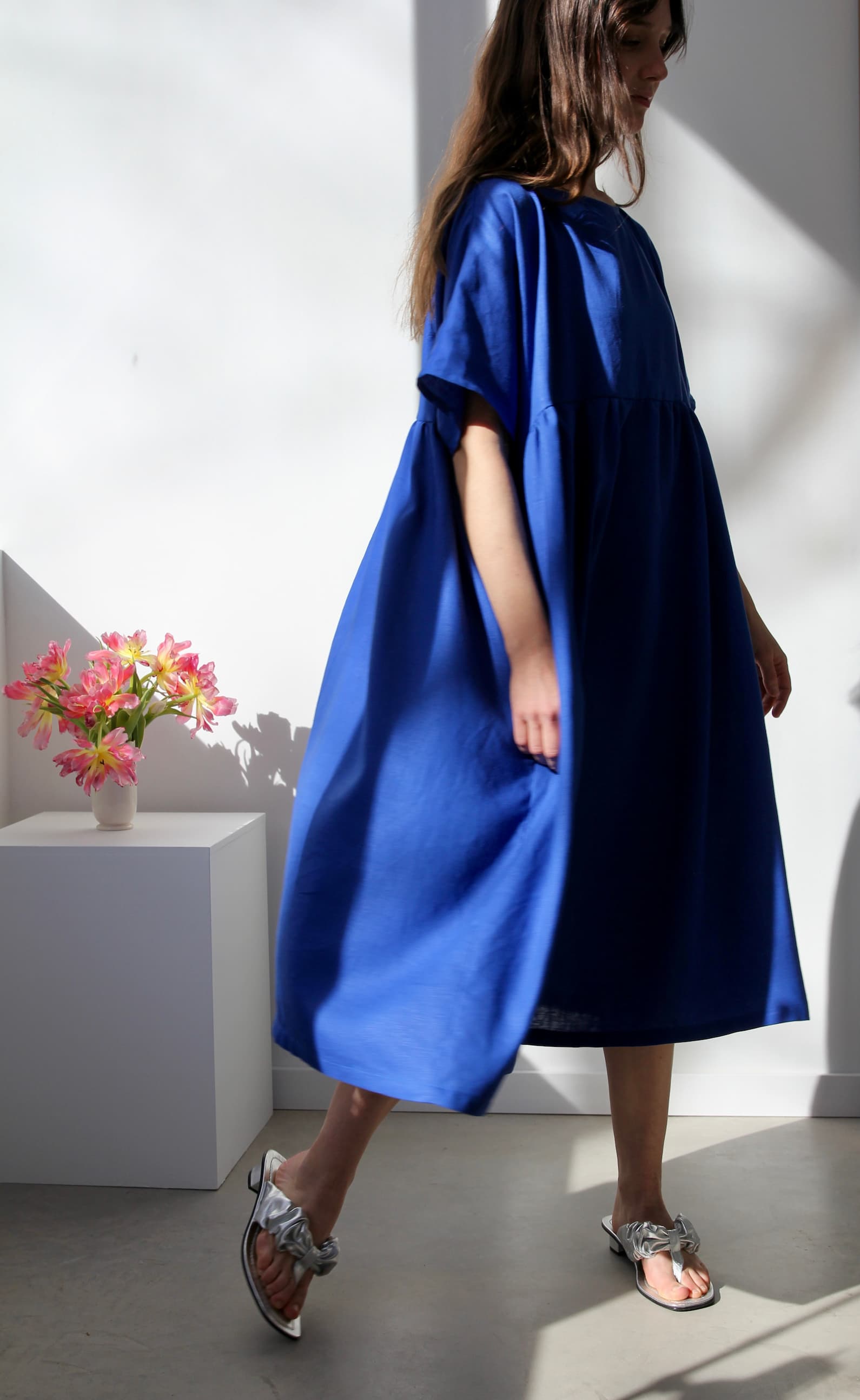 A person in a flowing Cécilia Classic Dress in Matisse Blue by Noémiah embodies relaxed femininity as they stand in a sunlit room, slightly turned away from the camera with their hair cascading over their shoulder. In the background, a small bouquet of pink flowers rests on a white pedestal.