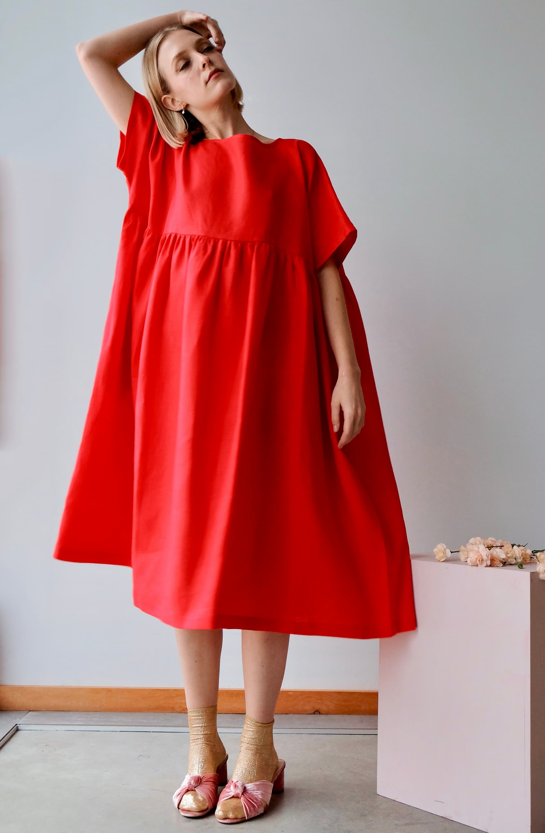 Woman wearing a vibrant red midi-length Noémiah Cécilia classic dress with a gathered waist, gold sparkly socks, and blush-pink heeled sandals.