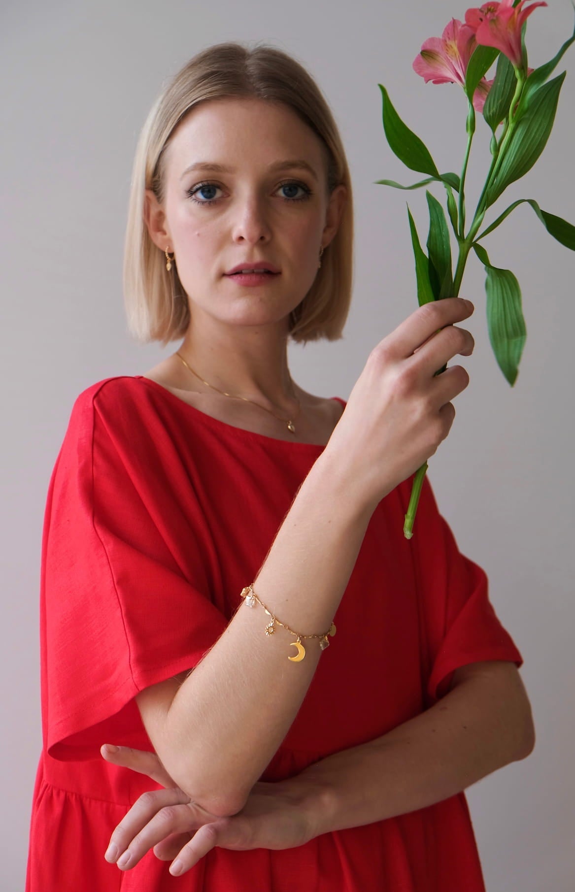 A woman in a limited edition red Cécilia Classic Dress by Noémiah holds a pink flower. She has short blonde hair and wears a gold bracelet with small charms, embodying relaxed femininity against a plain, light-colored background.