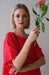 A woman in a limited edition red Cécilia Classic Dress by Noémiah holds a pink flower. She has short blonde hair and wears a gold bracelet with small charms, embodying relaxed femininity against a plain, light-colored background.