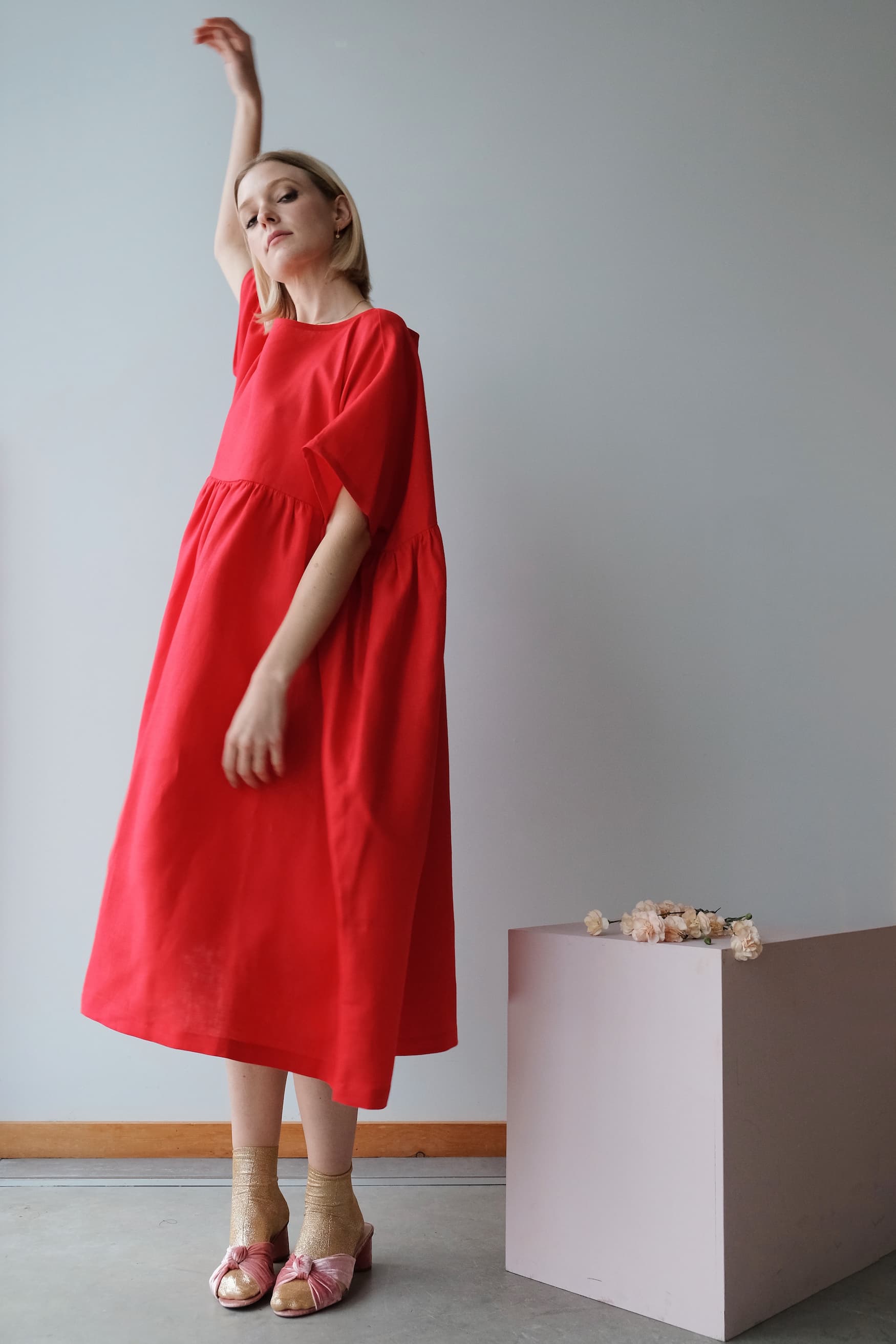 Wearing the limited edition, loose-fitting Cécilia Classic Dress in red by Noémiah, a person conveys relaxed femininity while raising one arm beside a pink pedestal adorned with flowers. Clad in pink heels, they stand against a plain gray wall accented by a wooden baseboard.