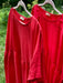 Two limited edition Cécilia Classic Dresses by Noémiah, made from soft, textured fabric, hang on wooden hangers outdoors. These red dresses boast long sleeves and a gathered waistline, exuding relaxed femininity amidst a lush greenery backdrop.