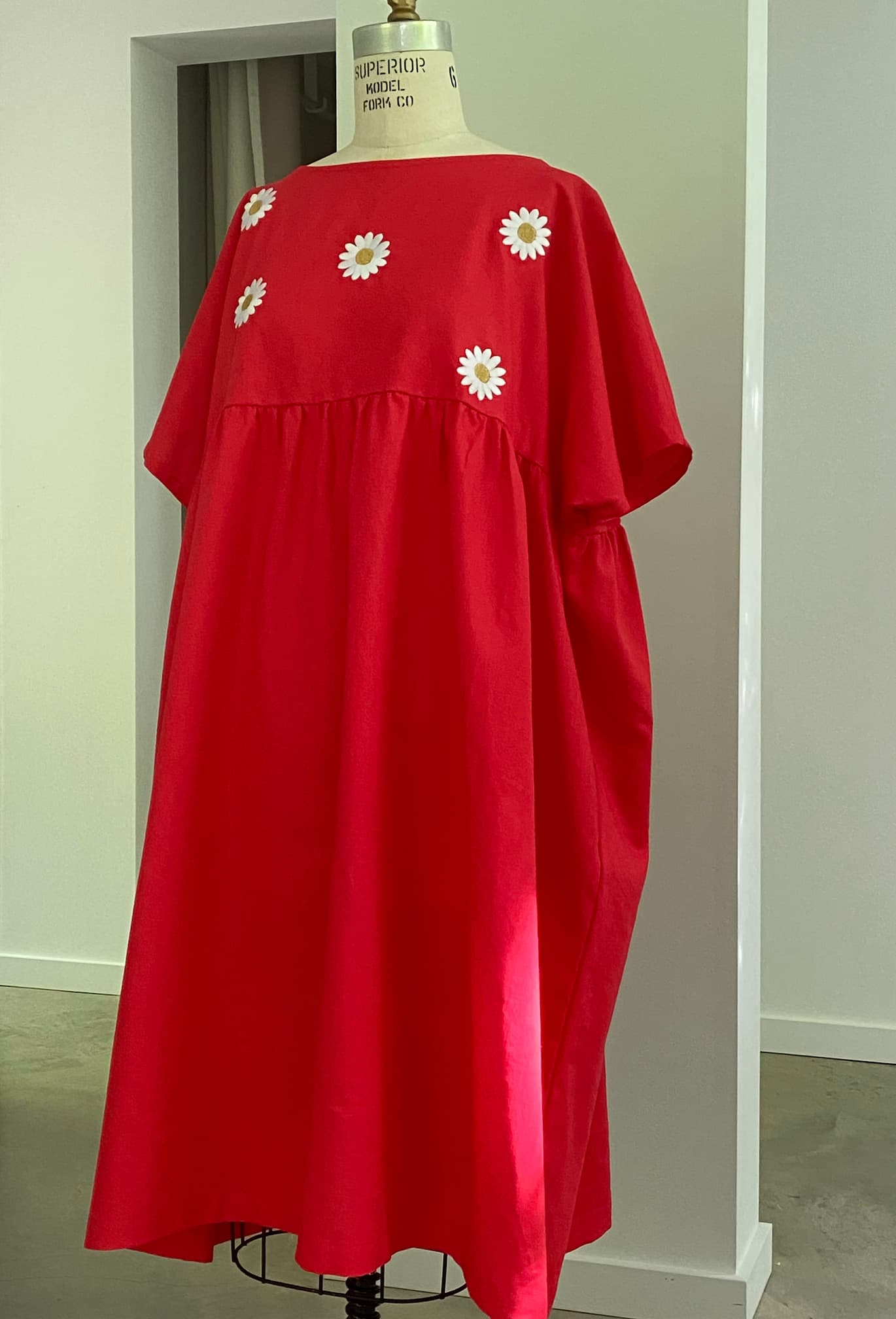 The limited edition Noémiah Cécilia Classic Dress in Poppy Red showcases short sleeves and a loose fit, adorned with white daisy appliqués on the upper front. Set against a minimalist studio with neutral walls and soft lighting, it represents relaxed femininity.