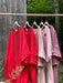 A row of five vibrant Noémiah dresses, featuring the limited edition Cécilia Classic Dress in red, is displayed on wooden hangers outside. These dresses range from bright red to soft pink, embodying a relaxed femininity against a rustic wooden wall and lush greenery backdrop.