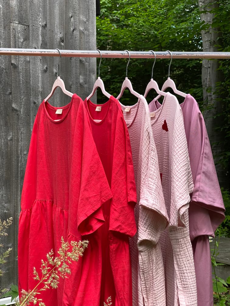 A row of five vibrant Noémiah dresses, featuring the limited edition Cécilia Classic Dress in red, is displayed on wooden hangers outside. These dresses range from bright red to soft pink, embodying a relaxed femininity against a rustic wooden wall and lush greenery backdrop.