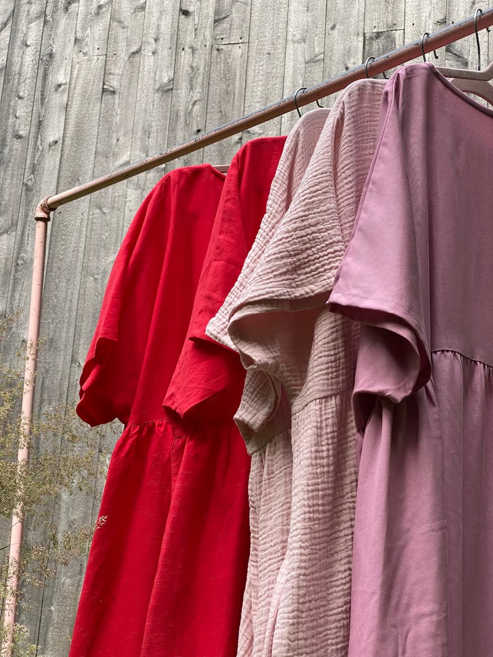 Three dresses on a pink metal rack against a wooden wall exude relaxed femininity, featuring the limited edition Cécilia Classic Dress in Poppy Red by Noémiah, joined by light and dusty pink dresses with varying textures and short sleeves.