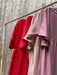 Three dresses on a pink metal rack against a wooden wall exude relaxed femininity, featuring the limited edition Cécilia Classic Dress in Poppy Red by Noémiah, joined by light and dusty pink dresses with varying textures and short sleeves.