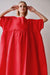 A person with short blond hair is wearing a Cécilia Classic Dress in red by Noémiah, standing with arms raised near their head against a plain background. The perspective from below highlights the voluminous shape of the dress, exuding an air of relaxed femininity.