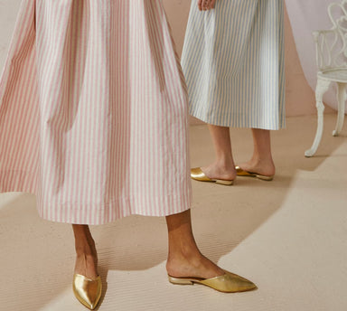 Two individuals don pastel-striped Cécilia dresses by Noémiah and gold pointed flats, creating an aura of relaxed femininity indoors. The space is adorned in soft hues, with the limited edition dress drawing attention and a white chair elegantly positioned in the background.