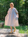 A person wearing the Cécilia Striped Dress by Noémiah and yellow shoes stands on a wooden deck, surrounded by lush green trees and plants, enjoying a sunny day with relaxed femininity.