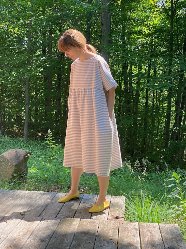 In a lush forest setting, an individual stands on a wooden platform wearing the loose-fitting Cécilia Striped Dress by Noémiah, paired with yellow shoes. Sunlight filters through the trees, casting gentle shadows on the ground and perfectly capturing the essence of relaxed femininity.