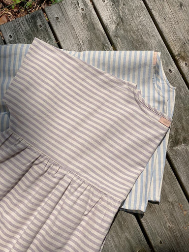 Two Cécilia Striped Dresses by Noémiah are laid on a wooden deck, embodying relaxed femininity. One dress features gray and white stripes, while the other showcases light blue and white stripes. Made from cotton canvas, these simple yet graceful designs are bathed in sunlight.