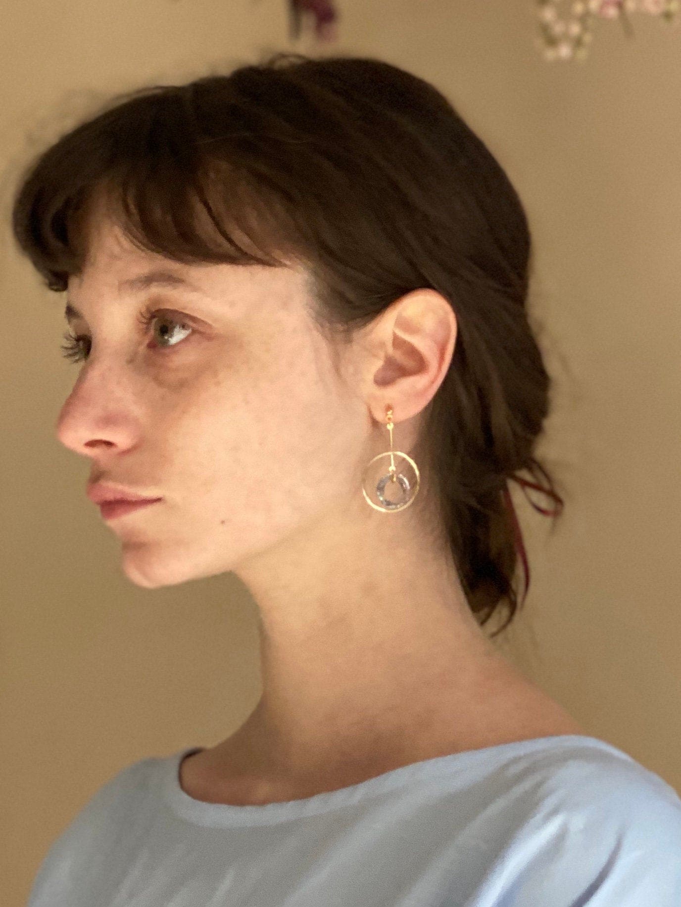 A woman with short brown hair is pictured from the side, wearing a light blue top and Céleste Earrings by Noémiah, which are adorned with Swarovski crystals for a bold everyday look. The background is softly lit and blurred, drawing attention to her thoughtful expression.