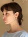 A woman with short brown hair is pictured from the side, wearing a light blue top and Céleste Earrings by Noémiah, which are adorned with Swarovski crystals for a bold everyday look. The background is softly lit and blurred, drawing attention to her thoughtful expression.