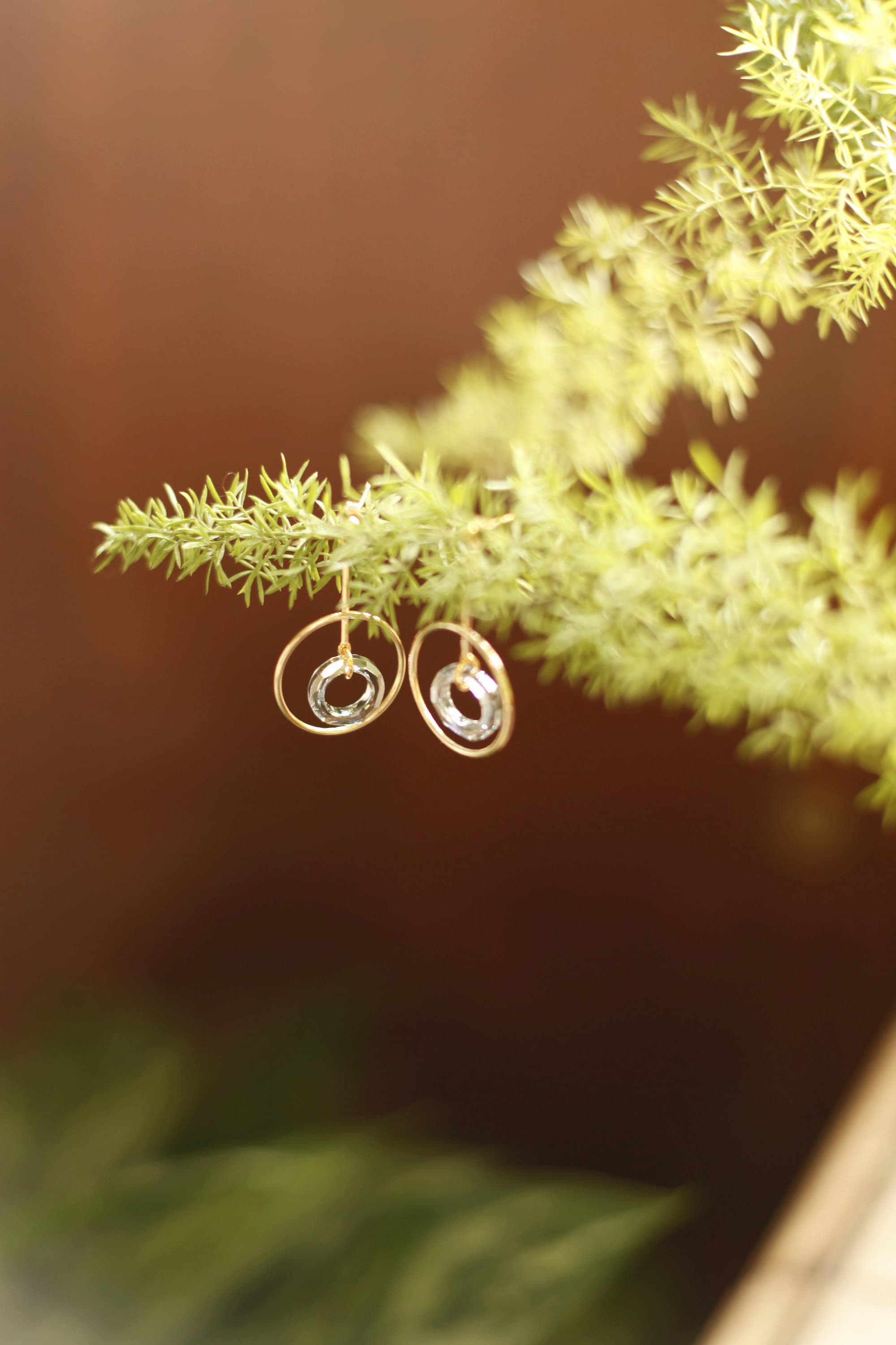 The Céleste Earrings by Noémiah, bold and designed for everyday wear, glisten with Swarovski crystals as they dangle from a green, leafy branch. The gold-plated hoops sparkle against a warm, blurred brown background, creating a natural and elegant setting.