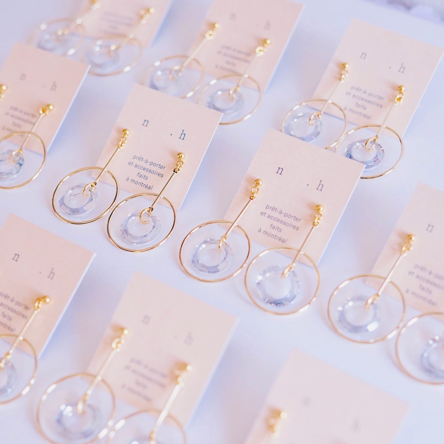 The Céleste Earrings by Noémiah, featuring gold-plated hoops adorned with clear circular pendants and enhanced with Swarovski crystals, are displayed on cards. These bold everyday earrings are neatly arranged in rows on a light-colored surface.