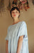 A person wearing Céleste Earrings by Noémiah and a light blue dress stands in a softly lit room. They have short, dark hair and are gazing directly at the camera. Dried flowers hang in the background, enhancing the serene and delicate atmosphere.