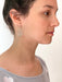 A woman with dark hair is shown in profile, wearing the Céleste Earrings by Noémiah, which are gold-plated and circular. Her light-colored top features subtle heart patterns, adding a touch of whimsy. The plain white background beautifully highlights her bold everyday earrings, making them glisten like Swarovski crystals.