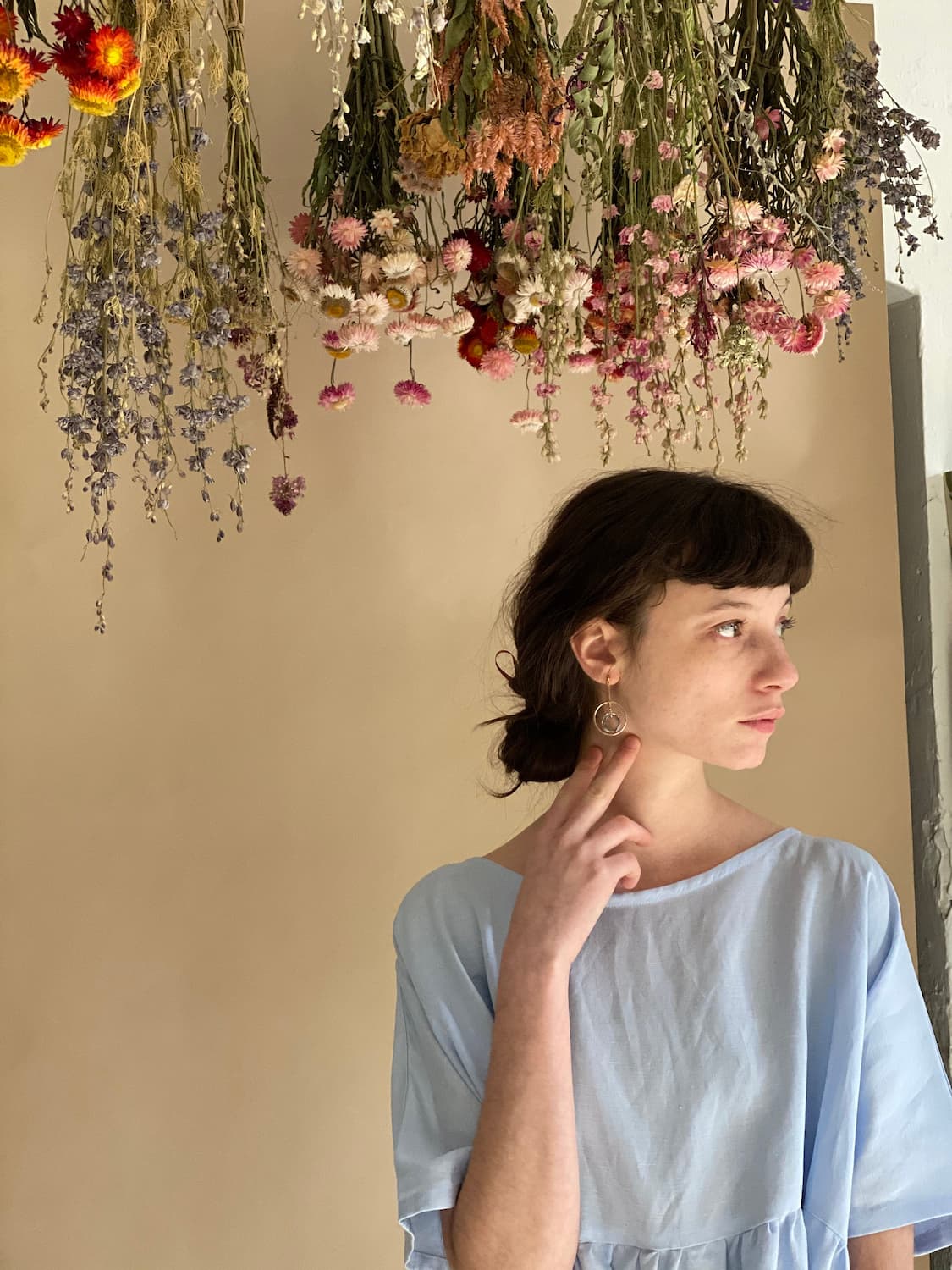 A person in a light blue dress gazes contemplatively to the side, standing beneath a display of colorful, hanging dried flowers against a neutral background, their Céleste Earrings by Noémiah catching the light with every slight movement.