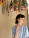 A person in a light blue dress gazes contemplatively to the side, standing beneath a display of colorful, hanging dried flowers against a neutral background, their Céleste Earrings by Noémiah catching the light with every slight movement.